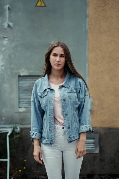 Молода красива дівчина з довгим волоссям у футболці та джинсовій куртці на фоні старовинної бетонної стіни . — стокове фото