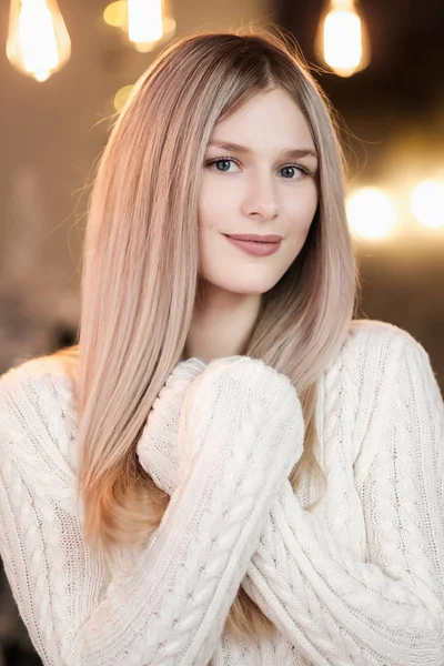 Молода красива блондинка в теплому білому в'язаному светрі на фоні домашньої затишної кімнати з різдвяними гірляндами . — стокове фото