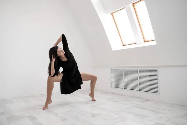 Молода жінка танцюрист в чорній сукні розтягування біля вікна. Білий зал, робітничий клас, йога асан. — стокове фото