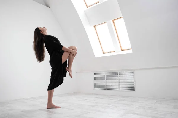 Una giovane ballerina vestita di nero si allunga vicino alla finestra. Sala bianca, la classe operaia, asana yoga . — Foto Stock