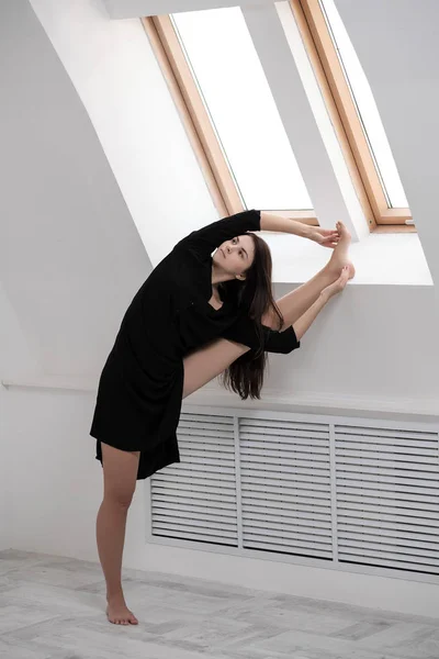 A young female dancer in a black dress is stretching by the window. White Hall, the working class, yoga asanas.