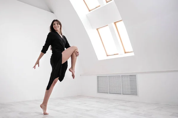 A young female dancer in a black dress is stretching by the window. White Hall, the working class, yoga asanas.