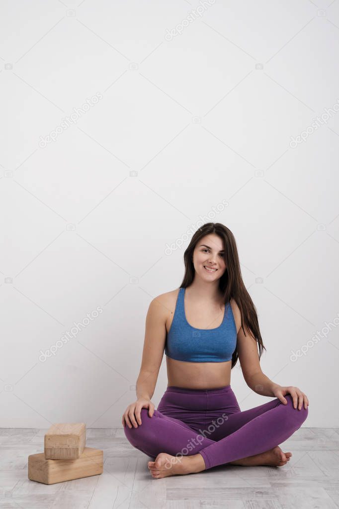 Young brunette woman in purple leggings and a sports t-shirt does exercises on the floor. Balances on the blocks and near the wall. White room.