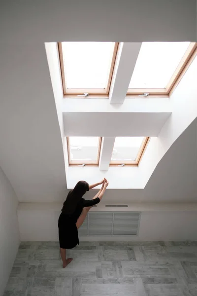 A young female dancer in a black dress is stretching by the window. White Hall, the working class, yoga asanas.