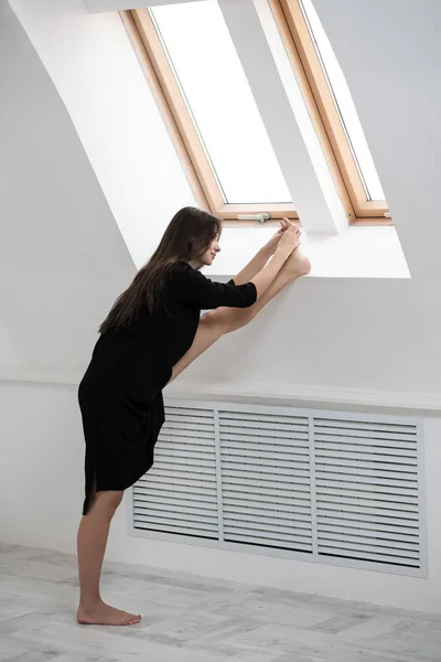 A young female dancer in a black dress is stretching by the window. White Hall, the working class, yoga asanas.