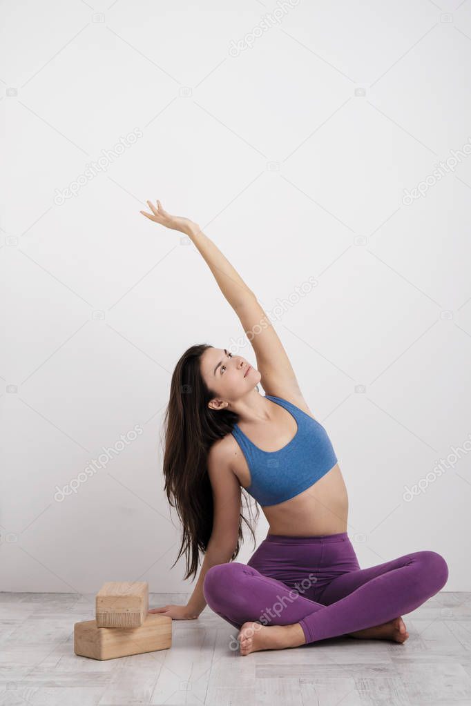 Young brunette woman in purple leggings and a sports t-shirt does exercises on the floor. Balances on the blocks and near the wall. White room.