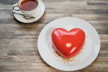 Kırmızı bir kalp şeklinde buzlu kek. Kahverengi ahşap masa ve çay arka planda.