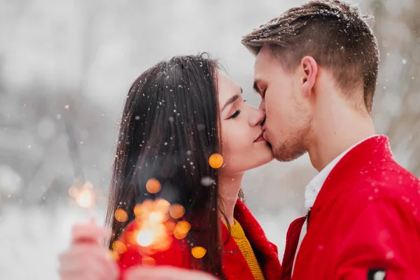 Young man and woman kiss in the winter in the park. Red clothes. In the hands of burning fireworks sparklers. Romantic fun date. — Stock Photo, Image