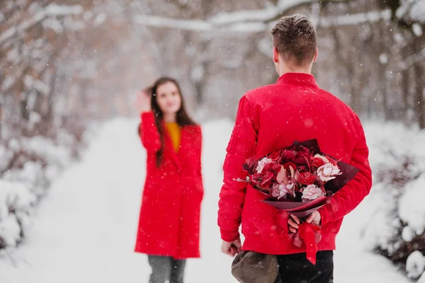 The man and the woman meet in snow-covered park in the winter. The guy is hiding behind a bouquet of flowers, a surprise for the girl. — Stock Photo, Image