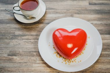 Kırmızı bir kalp şeklinde buzlu kek. Kahverengi ahşap masa ve çay arka planda.