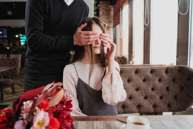 Bir genç adam kızı, karısı, gözlerini kapatır ve bir kafede cam kenarında bir buket kırmızı çiçek verir.