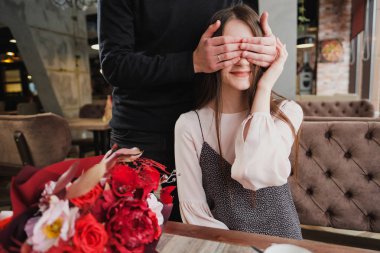 Bir genç adam kızı, karısı, gözlerini kapatır ve bir kafede cam kenarında bir buket kırmızı çiçek verir.
