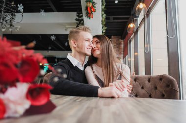 Genç çift sevgi, erkek ve kadın bir kafede romantik bir tarih. Pencerenin yanındaki bir masada oturmak. Masada bir buket kırmızı çiçek.
