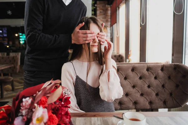 Bir genç adam kızı, karısı, gözlerini kapatır ve bir kafede cam kenarında bir buket kırmızı çiçek verir.