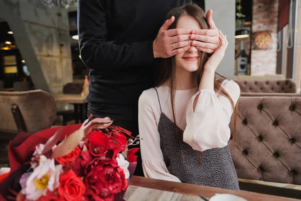 Bir genç adam kızı, karısı, gözlerini kapatır ve bir kafede cam kenarında bir buket kırmızı çiçek verir.