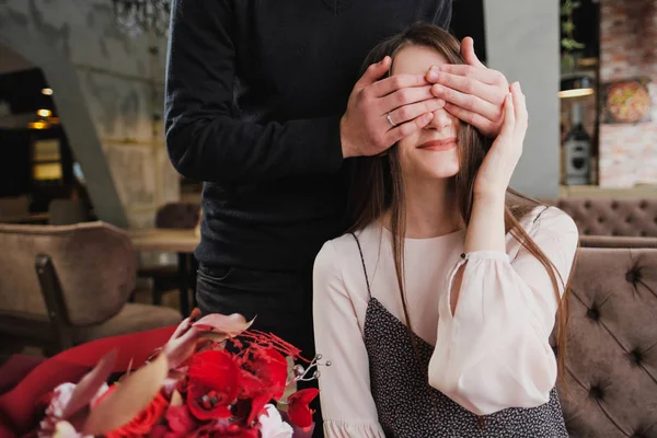 Un joven cierra los ojos a la niña, su esposa, y le da un ramo de flores rojas en un café junto a la ventana . —  Fotos de Stock