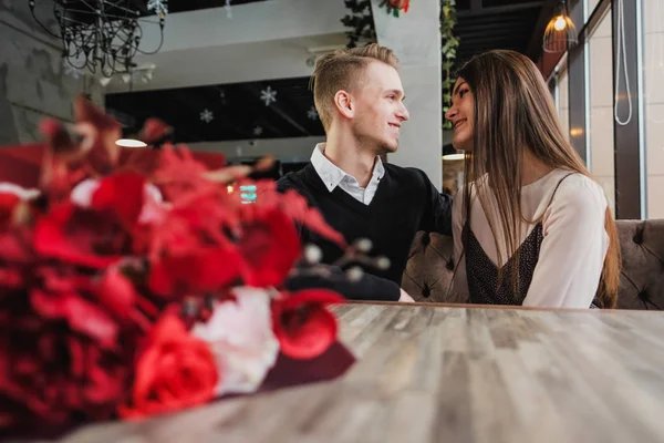 Mladý pár v lásce, muž a žena na romantické rande v kavárně. Sedět u stolu u okna. Na stole kytice červená. — Stock fotografie