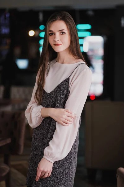 Ritratto di una giovane ragazza con i capelli lunghi sullo sfondo di segni luminosi all'interno di un caffè o ristorante. Abito e camicetta neri, eleganti e attraenti . — Foto Stock