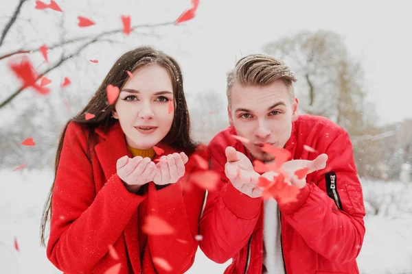 Молодая пара, парень и девушка, на улице зимой в красной одежде дуют на конфетти в виде сердец . — стоковое фото