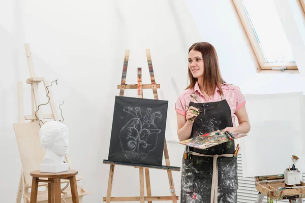 Młoda kobieta malarz w jasny biały studio rysuje obraz na płótnie na sztalugach. — Zdjęcie stockowe
