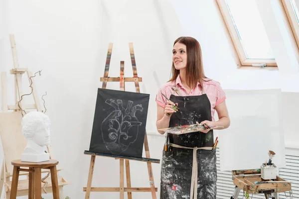 Uma jovem pintora em um estúdio branco brilhante desenha um quadro em tela em um cavalete . — Fotografia de Stock