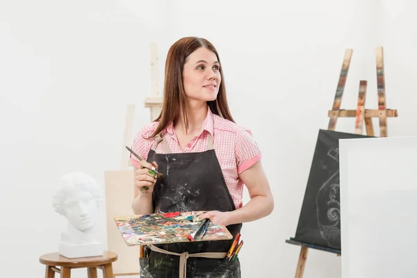 En ung kvinna målare i en ljusa vita studio ritar en bild på duk på ett staffli. — Stockfoto