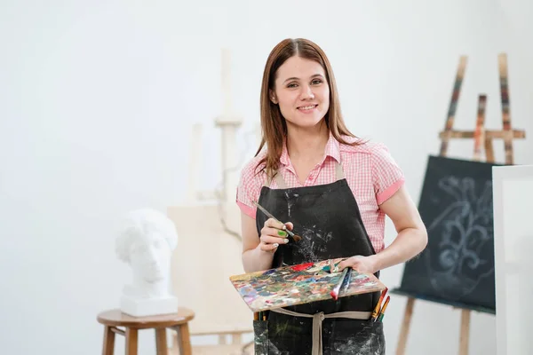 Una joven pintora en un estudio blanco brillante dibuja un cuadro sobre lienzo en un caballete . —  Fotos de Stock