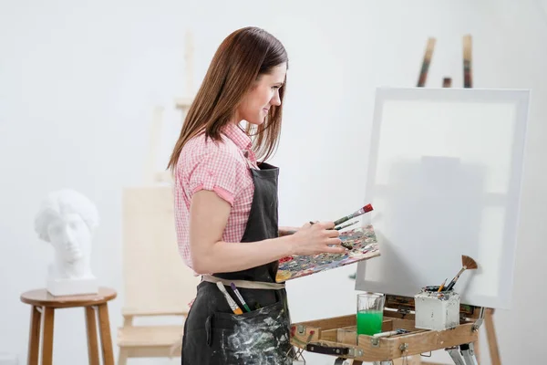 Uma jovem pintora em um estúdio branco brilhante desenha um quadro em tela em um cavalete . — Fotografia de Stock