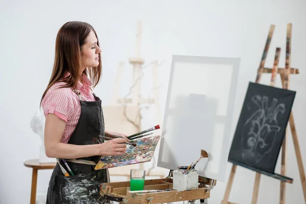 A young woman painter in a bright white studio draws a picture on canvas on an easel.