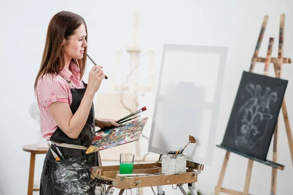 Una joven pintora en un estudio blanco brillante dibuja un cuadro sobre lienzo en un caballete . — Foto de Stock