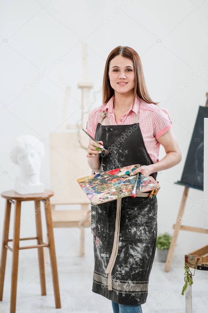 A young woman painter in a bright white studio draws a picture on canvas on an easel.