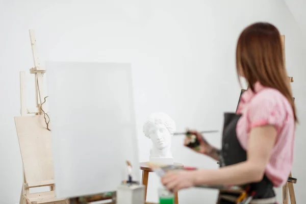 Joven hermosa niña pintor en un estudio blanco dibuja en un caballete sobre lienzo . — Foto de Stock