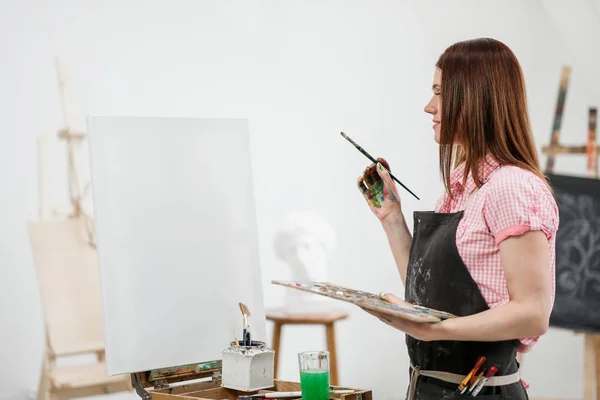 Jovem pintor bonito menina em um estúdio branco se baseia em um cavalete na tela . — Fotografia de Stock