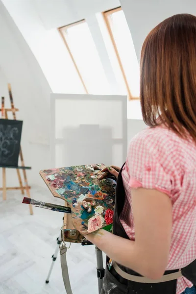 Tineri frumos fata pictor într-un studio alb desenează pe un șevalon pe pânză . — Fotografie, imagine de stoc