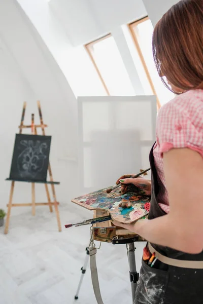 Joven hermosa niña pintor en un estudio blanco dibuja en un caballete sobre lienzo . —  Fotos de Stock