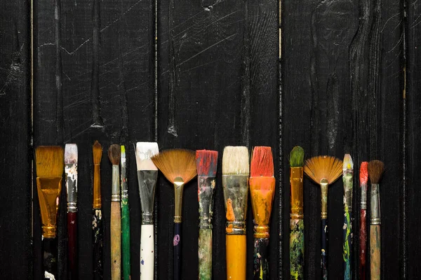 Artistas ferramentas, escovas em tinta estão em uma fileira em um fundo de madeira preta . — Fotografia de Stock