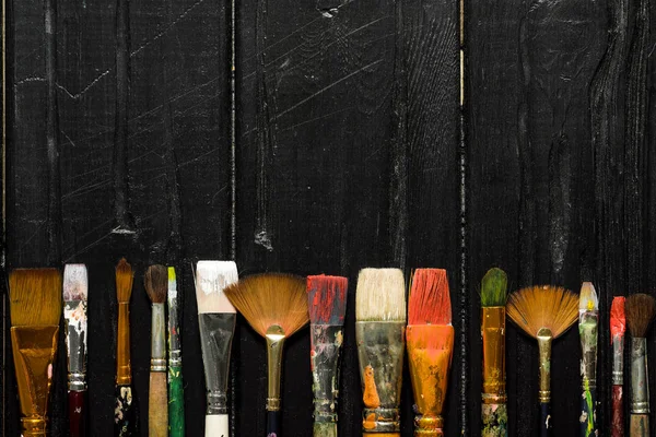 Artists tools, brushes in paint are in a row on a black wooden background. — Stock Photo, Image