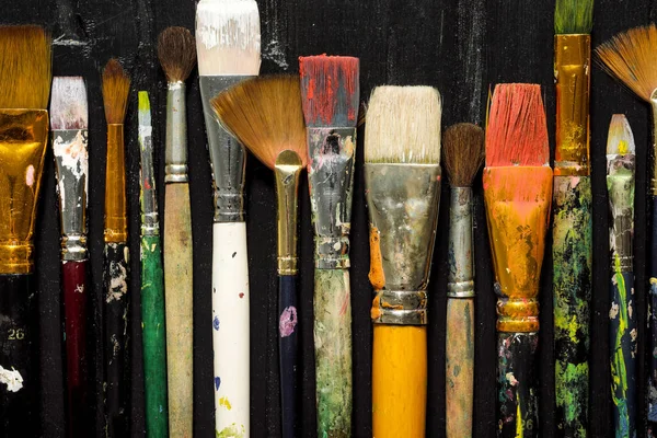 Herramientas de los artistas, pinceles en pintura están en una fila sobre un fondo de madera negro . — Foto de Stock