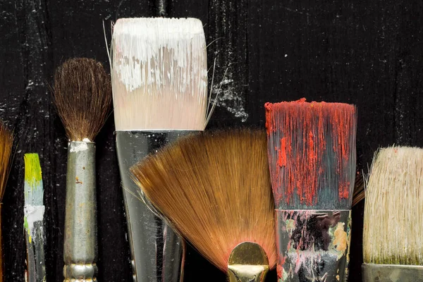Artistas ferramentas, escovas em tinta estão em uma fileira em um fundo de madeira preta . — Fotografia de Stock