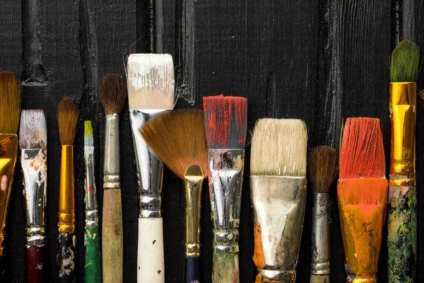 Artists tools, brushes in paint are in a row on a black wooden background.