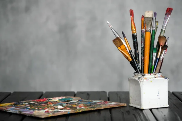 Gli strumenti di artisti, le spazzole, le vernici e una tavolozza si trovano su un tavolo di legno nero su uno sfondo di tessuto grigio . — Foto Stock