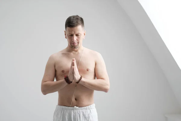 Un hombre vestido de blanco y con el pecho desnudo sobre un fondo blanco con las manos dobladas en el pecho. Meditación y oración, yoga . —  Fotos de Stock