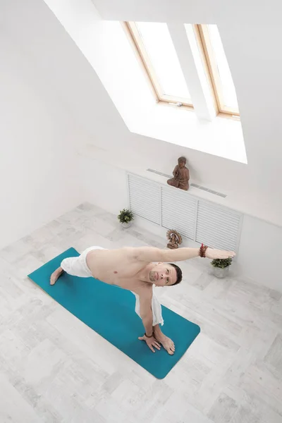 Un hombre con pantalones blancos y el pecho desnudo haciendo ejercicios. Yoga asanas y poses para estiramiento y meditación. Contra el telón de fondo de una brillante clase deportiva . — Foto de Stock