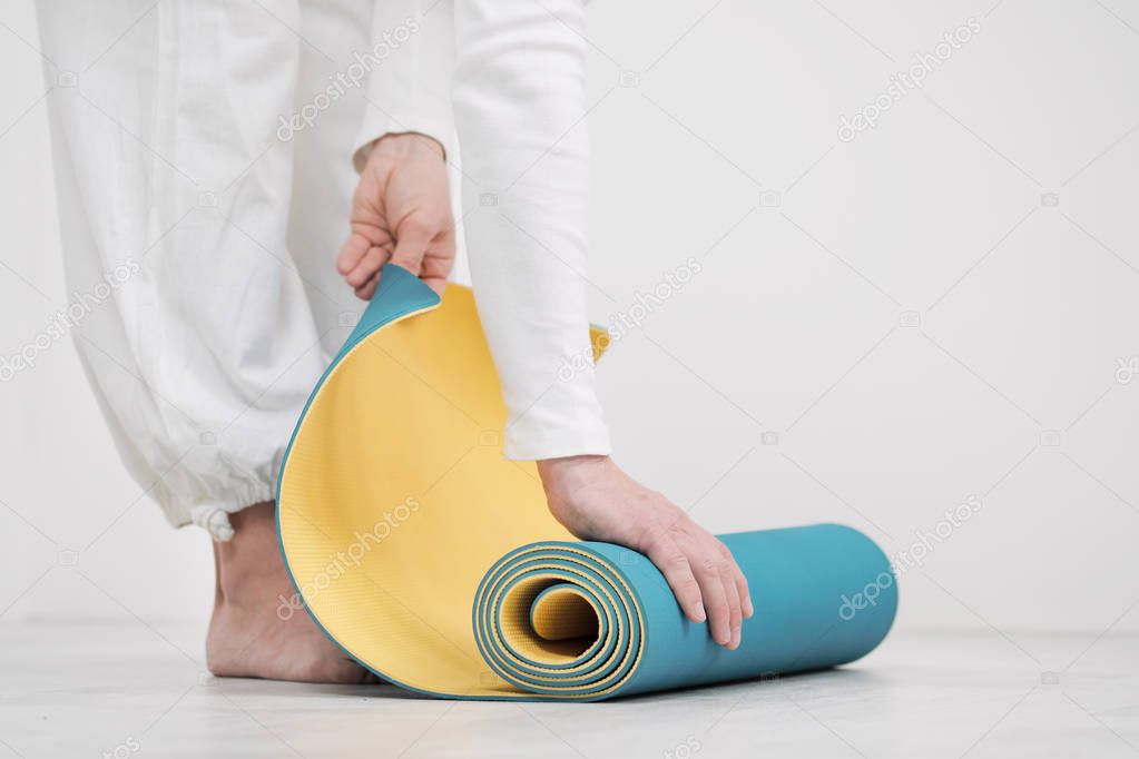A male teacher and trainer in white clothes are laying out a yellow and blue yoga mat, preparing for a fitness class.