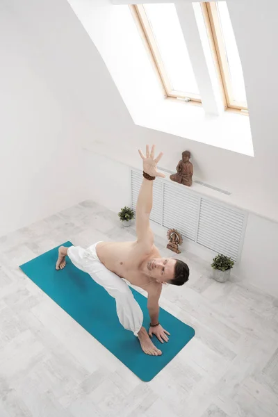 Un hombre con pantalones blancos y el pecho desnudo haciendo ejercicios. Yoga asanas y poses para estiramiento y meditación. Contra el telón de fondo de una brillante clase deportiva . — Foto de Stock