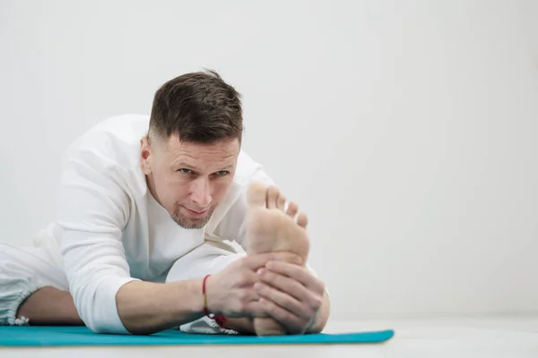 Handsome young man in white clothes does exercises. Yoga asanas and poses for stretching and meditating on a blue rug. Against the background of a white wall.