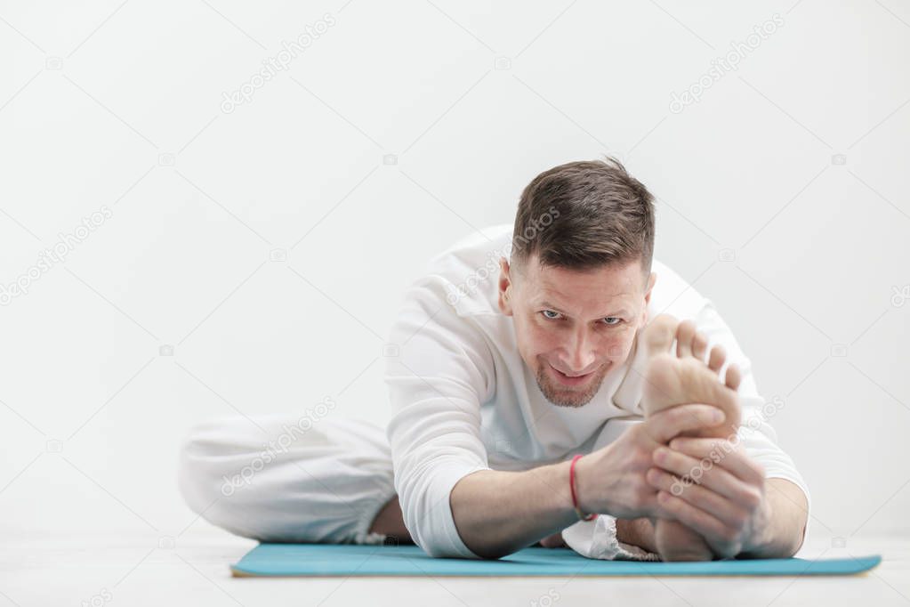 Handsome young man in white clothes does exercises. Yoga asanas and poses for stretching and meditating on a blue rug. Against the background of a white wall.