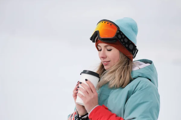 A beautiful young chewing woman in an extreme sports suit, hat and helmet with a mask on the winter slope is holding a paper cup with a hot coffee drink. — Stock Photo, Image