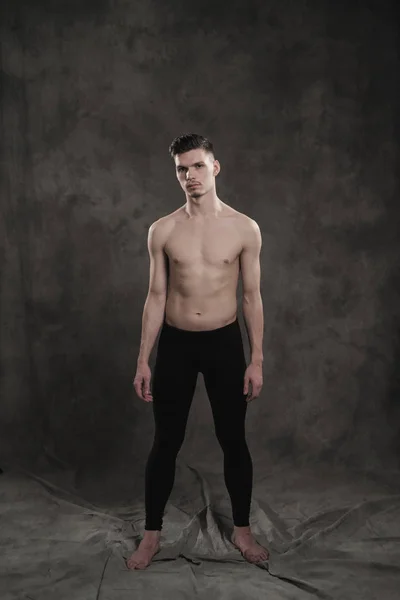 Una giovane ballerina di balletto maschile con leggings neri e un busto nudo esegue mosse di danza su uno sfondo grigio grunge, con una luce di luci e fumo . — Foto Stock