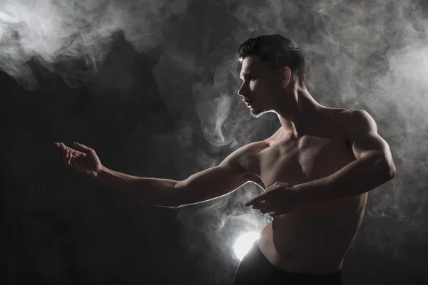 Un joven bailarín de ballet masculino con leggins negros y un torso desnudo realiza movimientos de baile sobre un fondo grunge gris, con una luz de luces y humo . —  Fotos de Stock